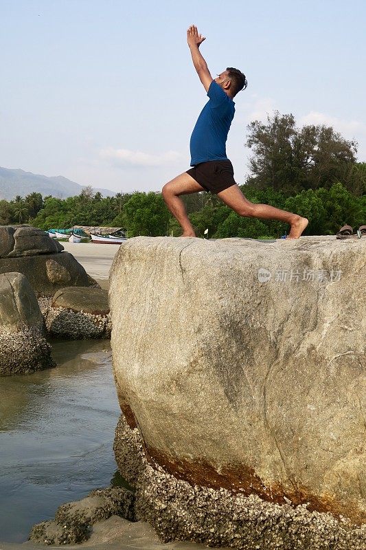 印度男子练习武士I姿势(Virabhadrasana I)瑜伽位置在岩石，水的边缘，帕洛伦海滩，果阿，印度